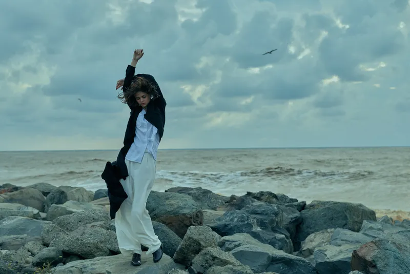 Woman standing on rocky shore by the sea, arms raised, wind catching flowing clothes, dramatic cloudy sky, coastal fashion mood on a rugged shoreline.