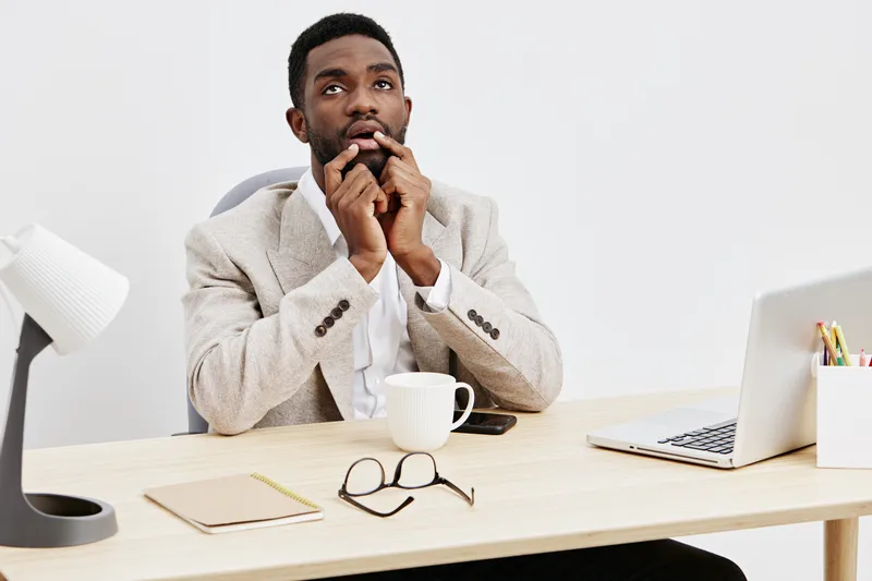 Thoughtful young man sitting at desk in office, holding his chin and contemplating, with laptop, notebook, glasses, and coffee mug on wooden table, professional workspace, business concept, neutral ba