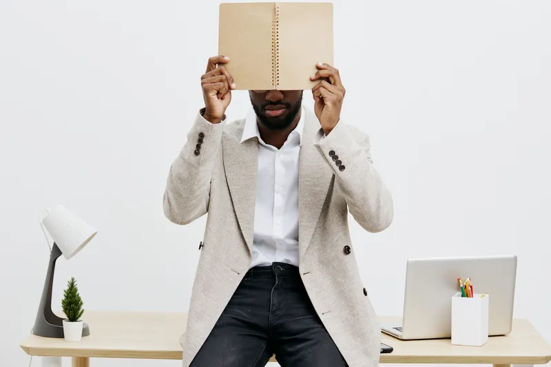 Stylish African-American man in formal blazer and white shirt sitting at modern office desk holding a blank notebook covering his face, creating a professional workspace scene with minimalist decor an
