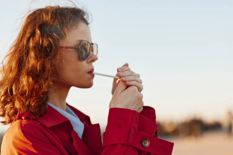 A woman with brown hair and a red jacket smokes a cigarette as the sun sets, creating a warm outdoor portrait with a contemplative mood and bright, soft light.