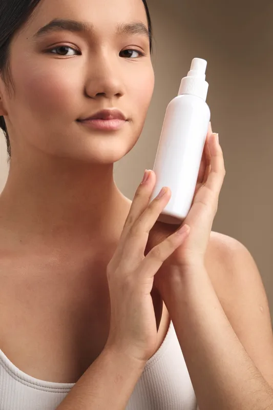 Asian woman holds a white skincare bottle near her face, showcasing a simple beauty routine in a soft studio setting that conveys calm confidence and clean aesthetics.