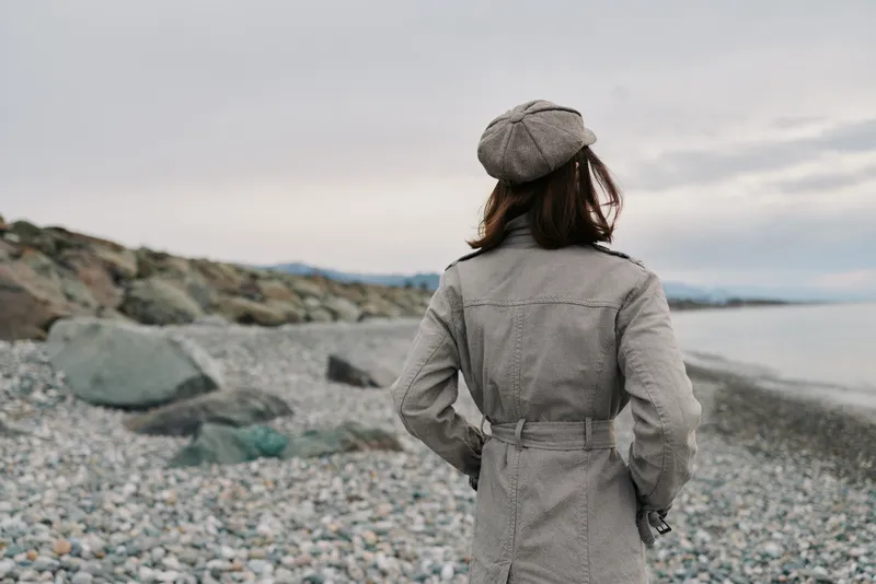 Woman wearing a checkered coat and hat stands on a rocky beach facing the sea under an overcast sky. Concept of autumn fashion, solitude, and coastal landscape.