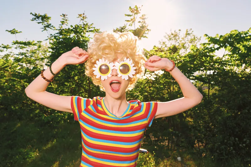 Woman outdoors in a sunny park, playful pose wearing a colorful striped shirt and sunflower glasses, retro film aesthetic, joyful portrait celebrating carefree lifestyle and creative fashion.