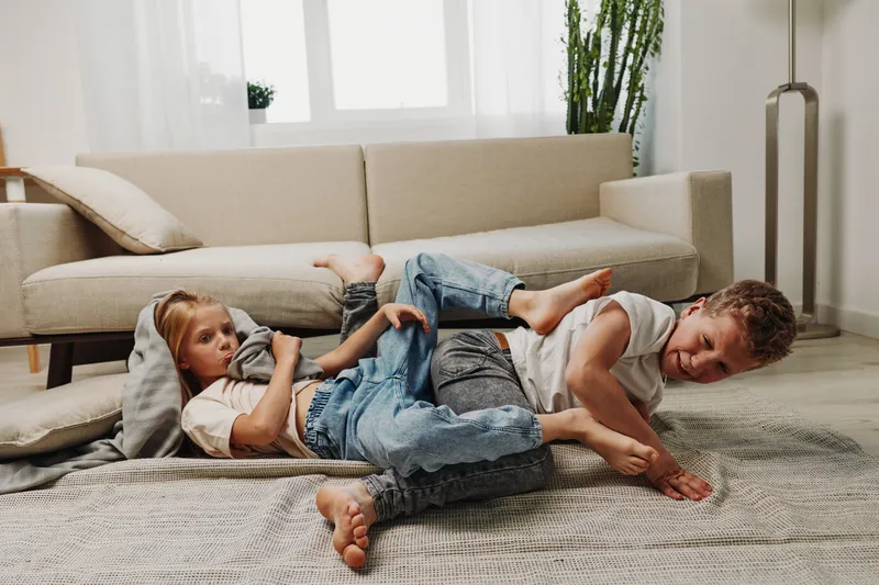 Two playful children in casual clothes wrestle on the carpet in a bright living room with modern sofa and plants. Concept of family fun and sibling bonding.