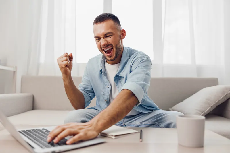 Excited man celebrating success while working on a laptop at home, showcasing modern lifestyle and self-motivation, bright and inviting atmosphere.