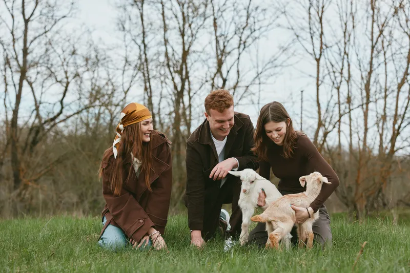 Lifestyle scene with three friends outdoors in a grassy field, interacting with playful goat kids, smiling and guiding the animals, capturing a warm candid moment of friendship and nature.