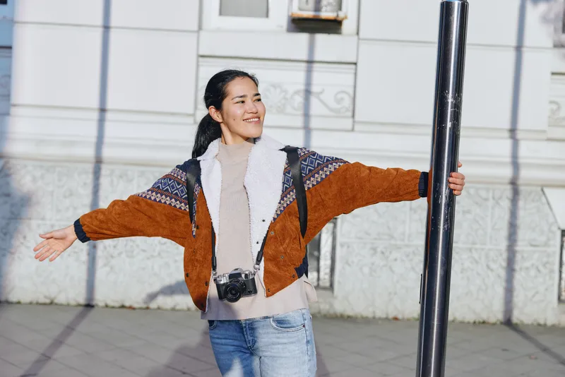 Woman outdoor street camera pose smile cardigan jacket scarf daylight, casual fashion urban streetwear happiness photography vibe, candid moment of joy and style on a sunny city street