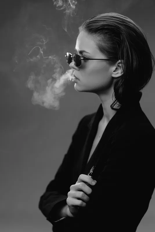 Black and white profile of a stylish woman wearing sunglasses exhaling smoke in a dark setting with moody lighting.
