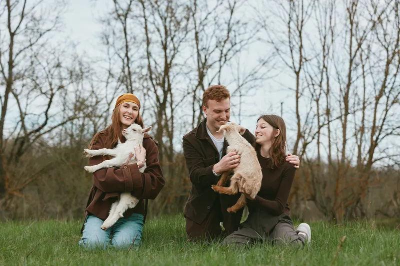 Happy group of friends outdoors with a playful goat kid, smiling and cuddling the animal, casual jackets in a grassy park, relaxed lifestyle moment captured in warm natural light.