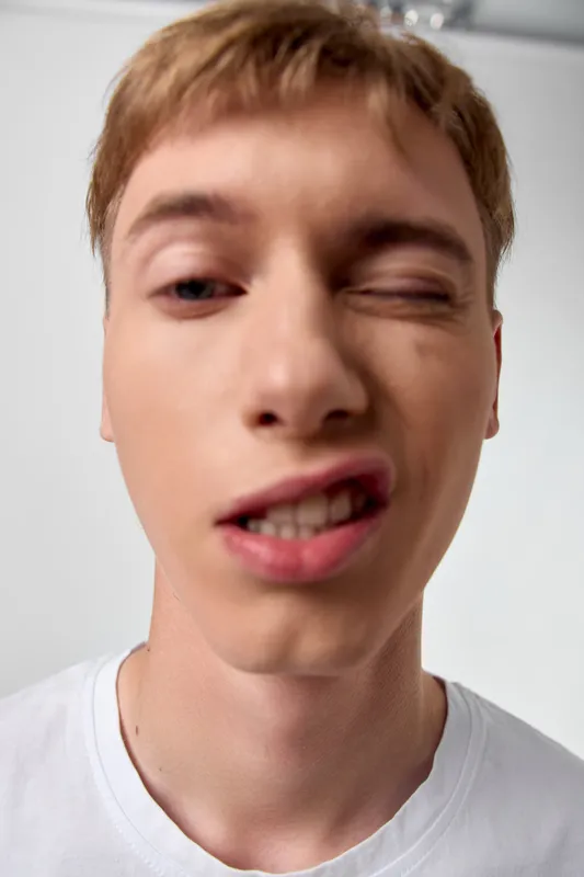 Close up of a young man making a playful face, one eye closed and lips twisted, wearing a white t shirt, casual studio shot conveying humor and spontaneity