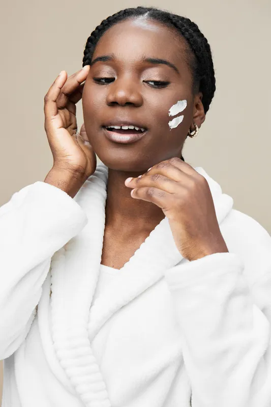 Young Black woman applying facial cream, wearing a cozy white robe, expressing joy in a soft, neutral background that highlights skincare and self care routines