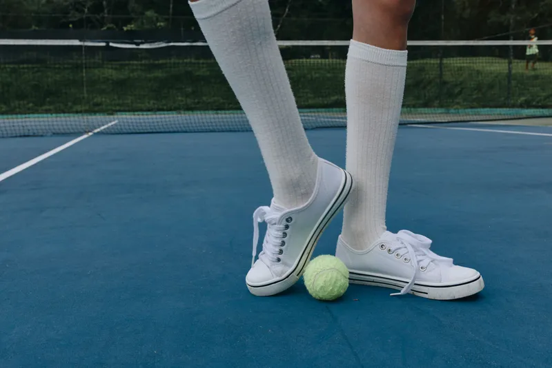 Young athlete in white tennis shoes and knee high socks standing with a tennis ball on a blue court, showcasing energy and a sporty lifestyle during summer