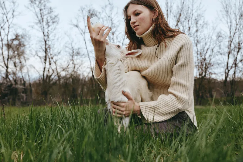 A woman in a beige knit sweater holds a goat kid in a grassy field, sharing a calm, intimate moment outdoors. Concept: rural lifestyle, animal bonding, and gentle care.