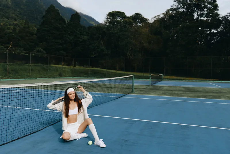 Young athletic woman in white sportswear sitting on a tennis court, showing confidence with a joyful expression, surrounded by greenery and blue tones, summer vibes