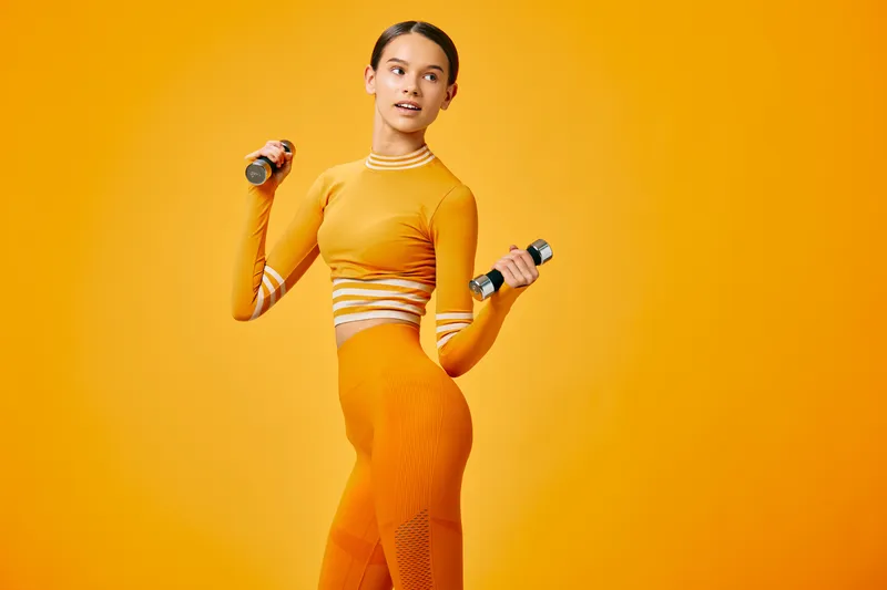 A fit woman in an orange athletic outfit lifts small dumbbells in a bold orange studio, showcasing strength, balance and confidence during a dynamic fitness routine.