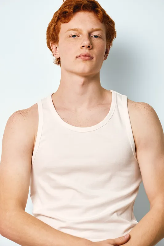Young man with red hair wearing white sleeveless shirt stands against light background with confident expression and relaxed posture showing casual style and natural look.