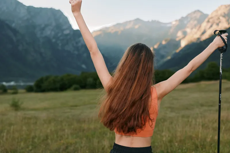 A young woman stands in a wide field with arms raised toward the sky, enjoying open nature. She exudes energy, confidence, and freedom in a vibrant outdoor scene.