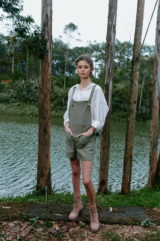 Stylish young model in green overalls and white blouse poses confidently near a serene lake, embodying a nature inspired fashion concept with a modern touch