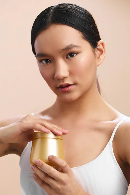 Asian woman holds a gold cosmetic jar and applies skincare cream, smiling softly in a clean studio setting, conveying calm beauty and modern self care.