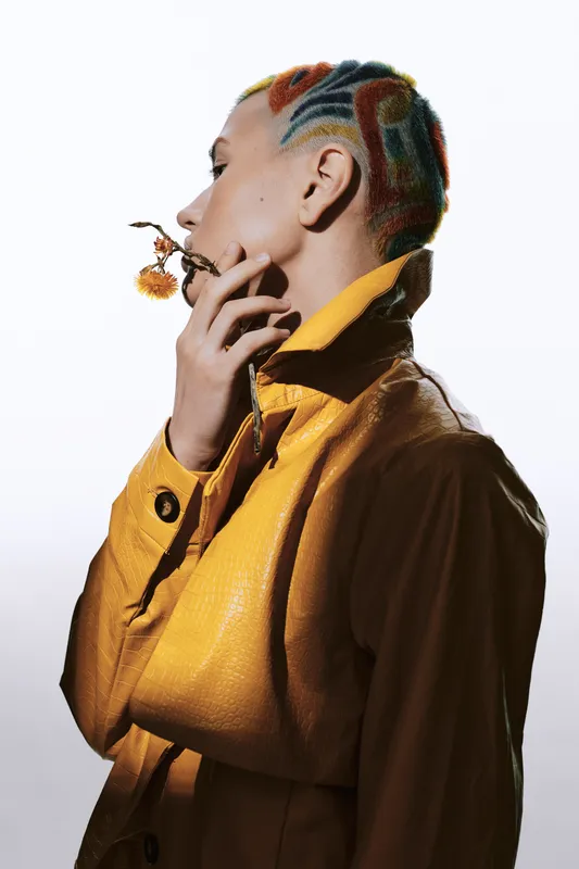 Profile of a woman with colorful braided hair wearing a yellow leather jacket, holding a small yellow flower thoughtfully against a bright background.