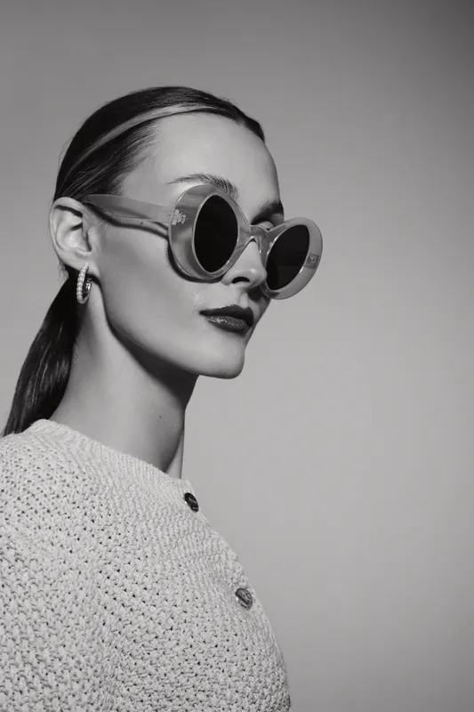 Portrait of a fair skinned Caucasian woman wearing round sunglasses and a textured sweater, styled hair pulled back, against a plain background. Fashion and elegance concept.