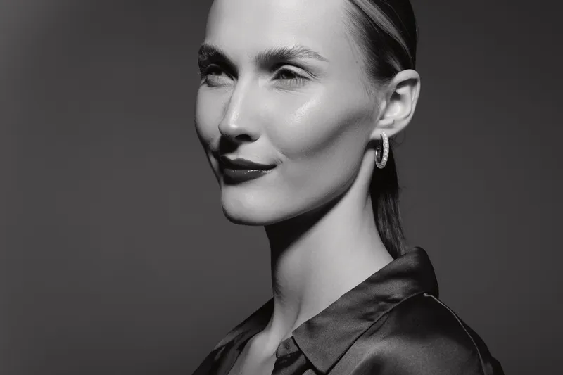 Elegant white woman with fair skin and slicked back hair wears hoop earrings and silk blouse, posing confidently on grey studio background. Concept of beauty, fashion, and portrait.