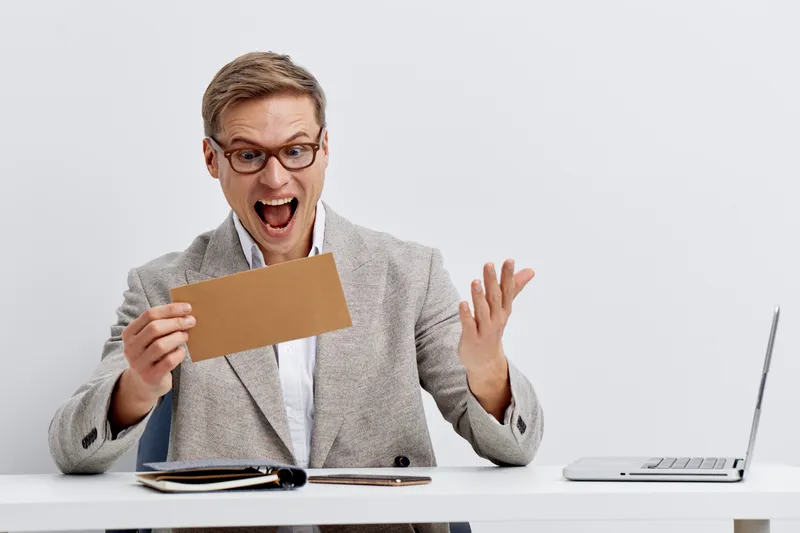 Happy excited middle-aged man in glasses holds an envelope and looks surprised at the camera while sitting at a desk with a laptop isolated on plain white background. People lifestyle concept
