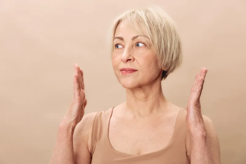 Mature woman with short blonde hair shows hand gesture, looking away with thoughtful expression. Neutral beige background emphasizes natural skin texture and casual style concept.