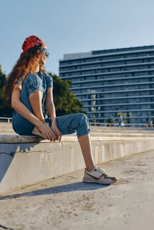 lifestyle woman in modern boho-western denim street style sitting outdoors with casual sneakers and red bandana, captured in warm film color tones on a sunny day