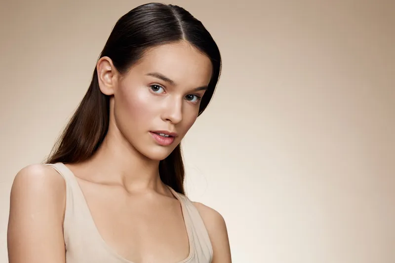 Elegant young woman with a natural look, showcasing healthy skin and subtle makeup against a soft, neutral background, representing beauty and confidence