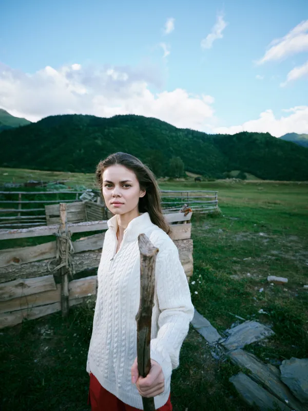 Woman in a white knit sweater holds an axe in a rural yard with a wooden fence, green hills and blue sky, portraying rugged outdoor life and rustic countryside atmosphere