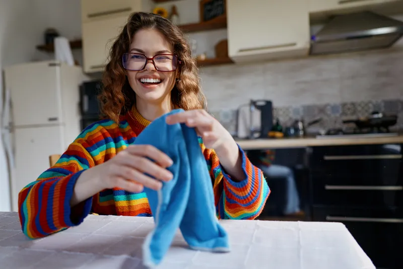 Smiling woman with curly hair wearing a colorful sweater folds a blue cloth at a kitchen table, conveying home chores, everyday life, and warmth with soft indoor lighting.