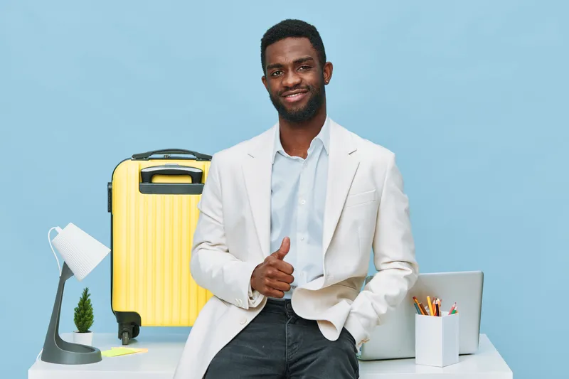 Smiling man with dark skin wearing light-colored blazer and shirt standing at modern travel desk with yellow suitcase, laptop, and office supplies on light blue background studio shot. Business travel