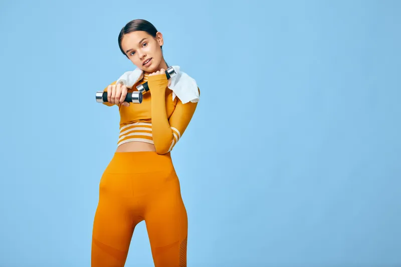 Fitness woman in orange athletic wear lifts a dumbbell with a towel over her shoulder, posing for a workout concept against a blue background, highlighting strength, exercise, training, and active
