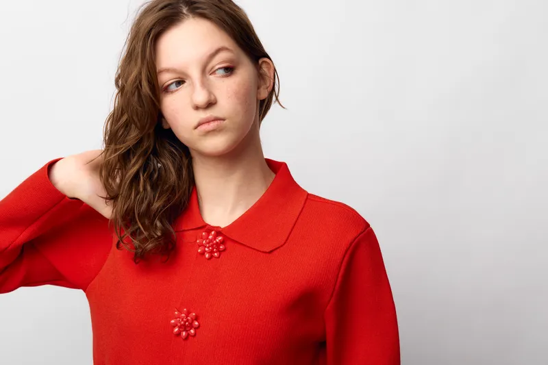 Young woman in red sweater looks away with thoughtful expression, hand touching hair. Studio portrait on white background. Concept of contemplation, mood, and casual fashion.
