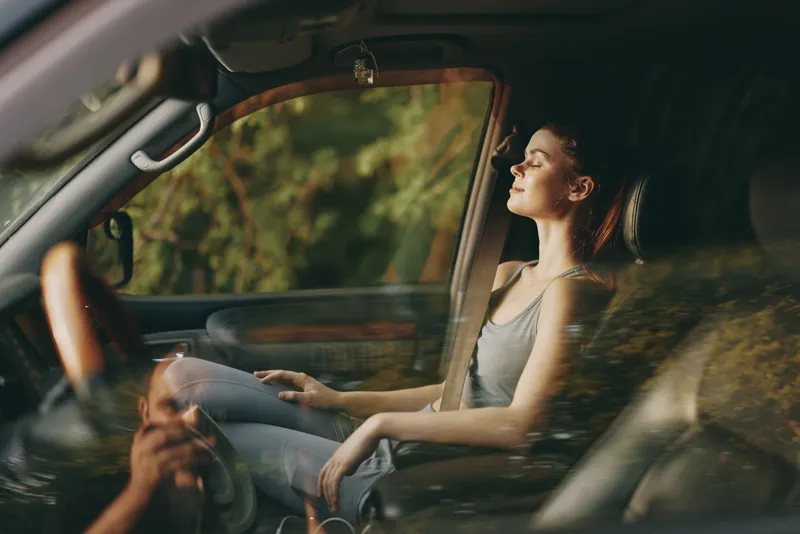 Young woman relaxing in a car, enjoying nature, with eyes closed, wearing casual gray outfit, surrounded by greenery, conveying peace and tranquility