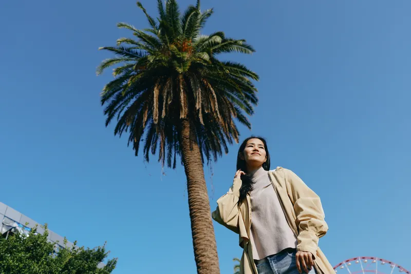 Palm tree outdoor scene with a woman in casual fashion posing under a bright sky in a sunlit park, capturing a summertime lifestyle moment in urban greenery
