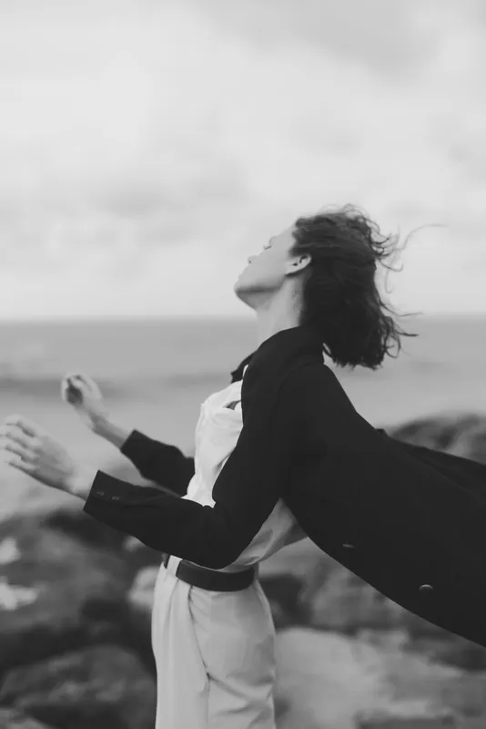 Wind swept figure stands along a rugged coast in a flowing coat, hair lifting, cape fluttering, capturing fashion and freedom in a cinematic monochrome scene.