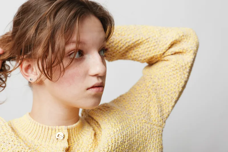 Young woman in yellow knitted sweater looks to side with thoughtful expression, hands behind head. Studio portrait on light background. Concept of contemplation, casual style, and natural beauty.