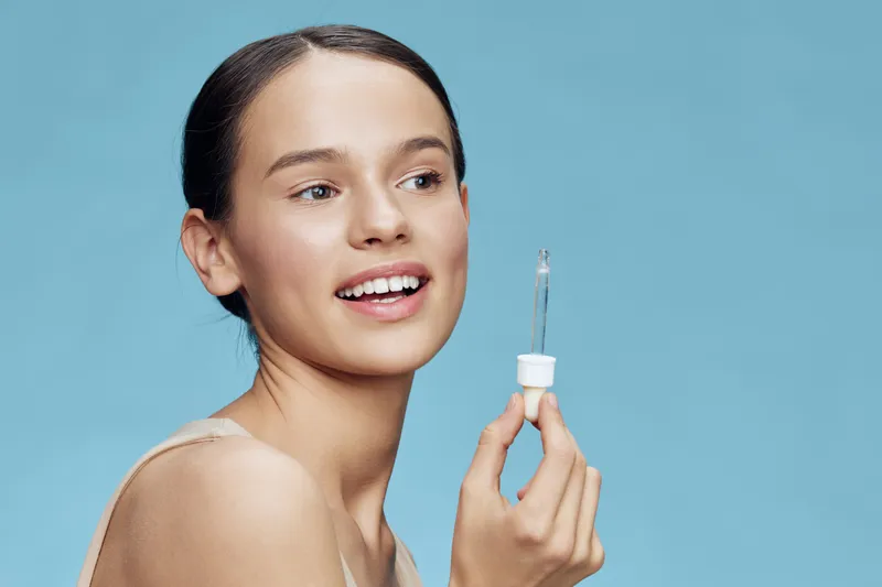 Young woman with clear skin holding a dropper bottle on a blue background, promoting skincare and healthy beauty concepts in a vibrant and fresh style