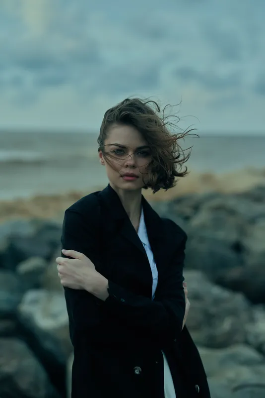 Woman in a dark blazer stands among rocky coast, wind in hair, moody ocean backdrop, cinematic style and thoughtful expression