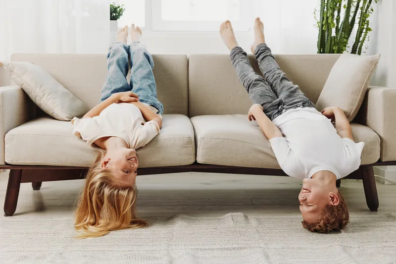Two happy children lie upside down on a beige couch in a bright living room, smiling and enjoying playful moments together.