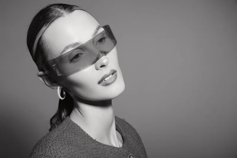 Portrait of a young white woman wearing futuristic sunglasses and hoop earrings. She poses with a neutral expression against a plain background. Fashion concept with minimalistic style.
