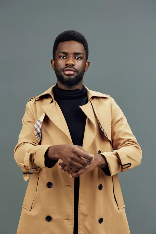 Stylish young African man in a beige trench coat posing confidently against a gray background, showcasing modern fashion and self assurance