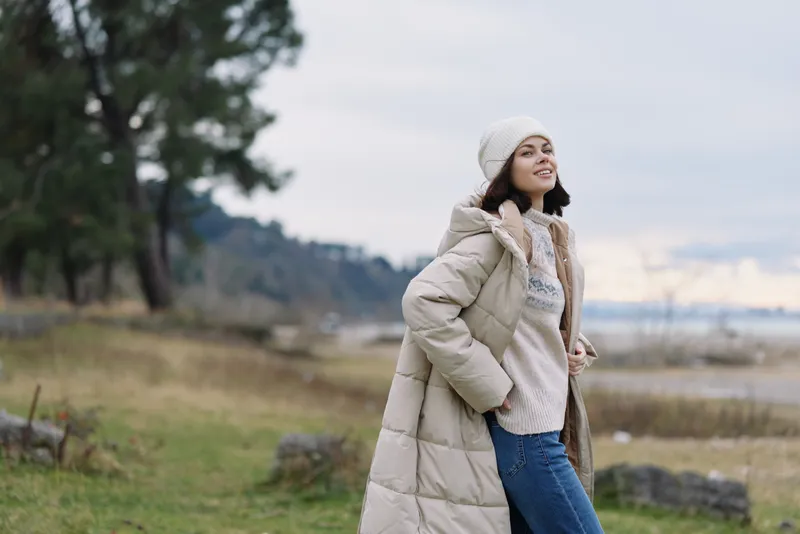 Woman in a long beige coat, scarf and beanie stands in a windy outdoor landscape by the lake, showcasing casual winter fashion, a serene sky and relaxed mood.