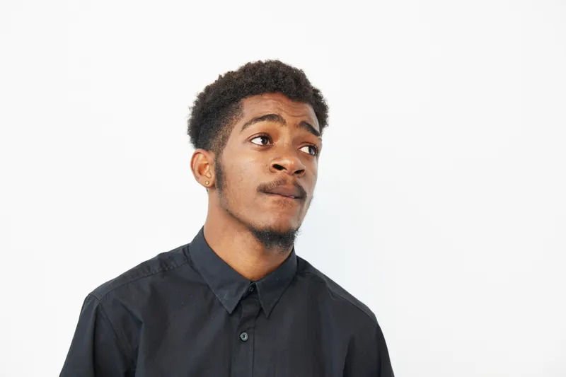 Black man looking thoughtfully into the distance, wearing a black shirt. The image conveys a sense of contemplation, style, and introspection.