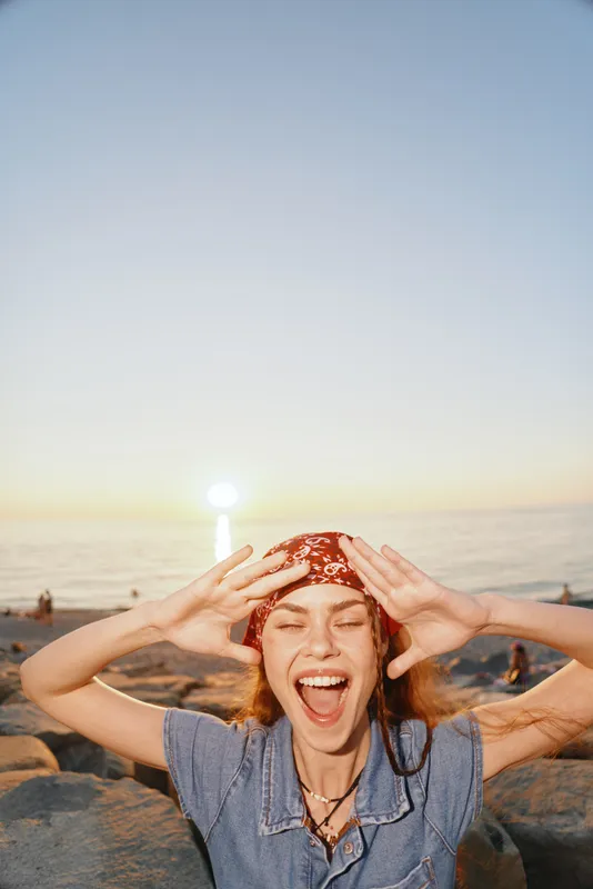 happy woman enjoying beach sunset with joyful expression, red headscarf and casual blue outfit in warm golden light