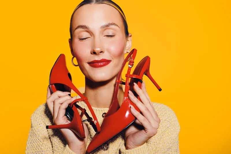 Happy woman with closed eyes holds red high heel shoes near face on bright yellow background. Concept of fashion, style, beauty, and joyful emotions in studio portrait.