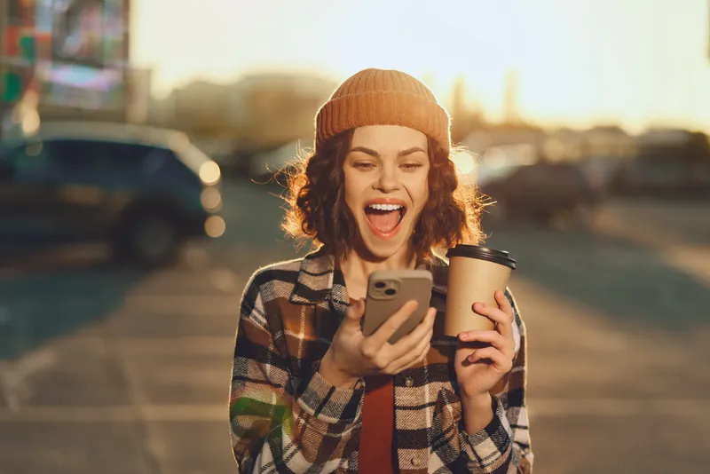 Woman holding coffee and phone on urban street, smile and beanie in golden hour glow. Candid lifestyle portrait with authenticity, mindful living and emotional storytelling mood.