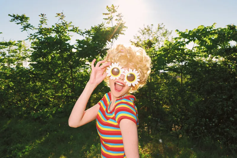 A stylish woman in a striped top and sunflower glasses enjoys a sunny outdoor moment, embodying retro film color and a carefree lifestyle. Concept for happiness, playful fashion, outdoor portrait.